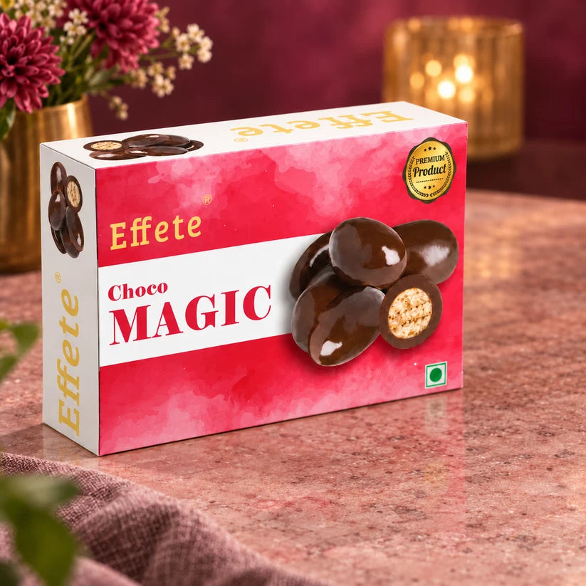 A white and red box of Effete Choco Magic chocolates sits on a table with flowers and a candle.