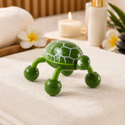 A green turtle massage tool rests on a soft white towel, surrounded by spa items like a candle and flowers.