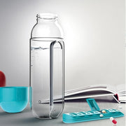 Clear glass bottle without its cap, next to a detached blue pill organizer and a blue cup on a desk.