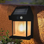 A black solar wall lamp with a clear light bulb, affixed to a rough stone wall with green leaves nearby.