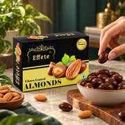 A hand reaches for a dark chocolate-covered almond from a white bowl, with a black and yellow box behind it.