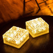 Two square solar lights with textured glass tops glow warmly on a dark, reflective table surface.
