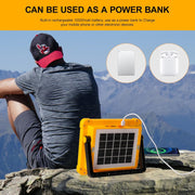 Person sitting on a rock in a mountainous landscape, charging a phone with the yellow solar floodlight.