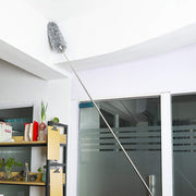 A person using the fully extended duster to clean a high corner of a white ceiling in a room with shelves and glass doors.