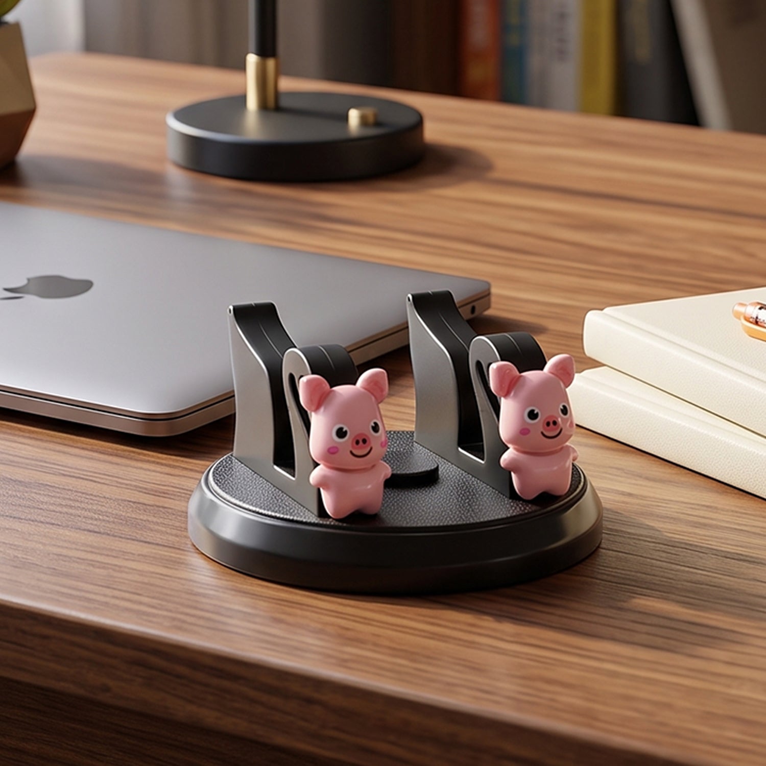A black phone holder with two pink pig figurines sitting on a wooden desk next to a laptop and books.