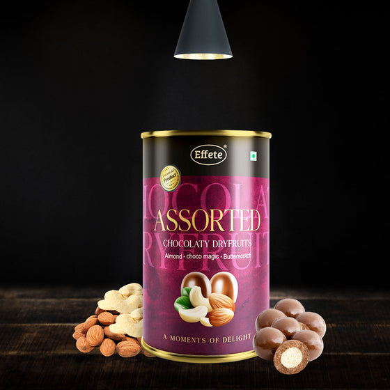 A tall, dark purple and black can of Effete Assorted Chocolaty Dryfruits sits on a wooden surface with loose almonds and