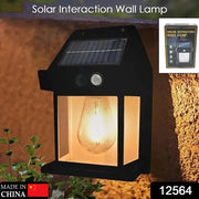 A black solar wall lamp with a clear bulb glowing warmly, mounted on a textured brown wall beside green plants.