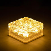 One square solar light with a textured glass top emits a warm yellow glow in darkness.