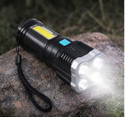 The black and silver flashlight with four illuminated LEDs rests on a textured rock outdoors, casting light forward.