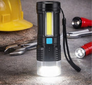 The black and silver flashlight stands upright on a concrete surface, with its base glowing brightly, surrounded by tools.