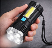 A hand holding the black and silver flashlight, with all four front LEDs illuminated and a side COB light visible.