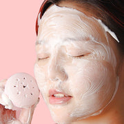 A woman with a foamy face mask uses a pink silicone facial cleanser to apply the product.