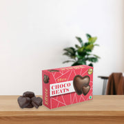 A red box of Effete Choco Beats heart-shaped chocolates sits on a wooden table with three dark candies beside it.