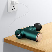 Teal massage gun with a round head, charging via a white cable plugged into a wall outlet.