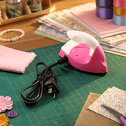 A person's hand guides a pink and white mini iron across a piece of light fabric on a green mat, with a power strip and