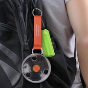 A clear circular case with an orange button and an orange strap is clipped to a dark backpack, with a green item also