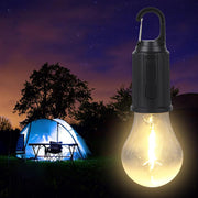 A glowing light bulb with a black carabiner hook hangs in front of a lit blue tent at night.