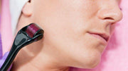 A person's face is shown in profile as a black derma roller is used on their cheek, with a pink towel visible.